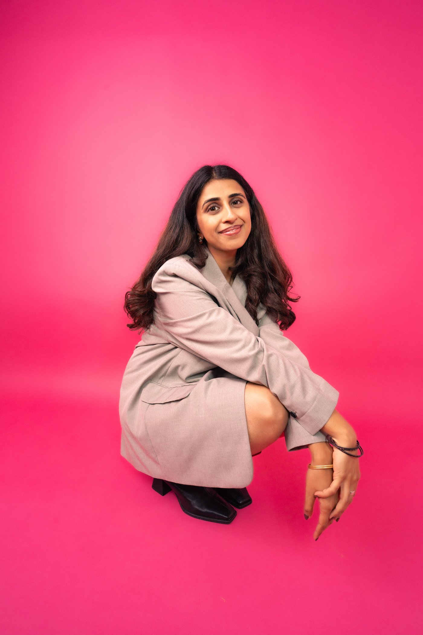 Ashna Yakoob crouching and smiling, wearing a taupe blazer, against a hot pink backdrop.