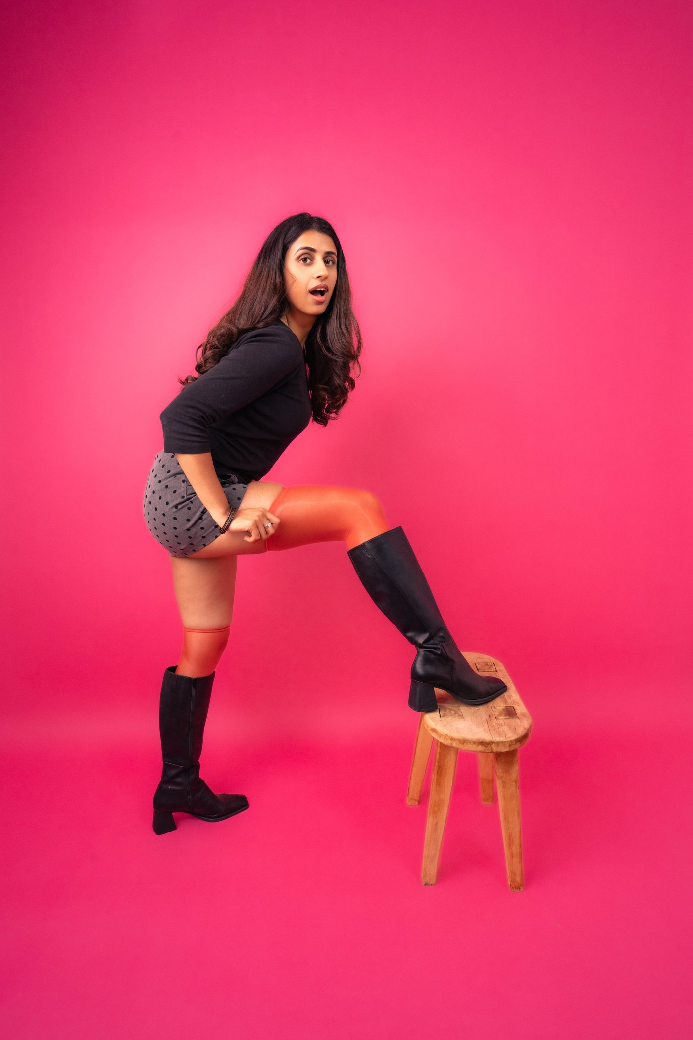 Ashna mid-bit, one boot up on a stool, mouth open mid-punchline.